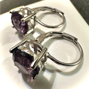 K&ouml;nnte beinhalten: Nahaufnahme von zwei Ohrringen mit B&uuml;gelverschluss. Jeder Ohrring hat einen gro&szlig;en, facettierten, lila Edelstein, der in einem silberfarbenen Metall gefasst ist. Die Ohrringe werden im Detail gezeigt, wobei die Farbe des Edelsteins und der Glanz des Metalls hervorgehoben werden.