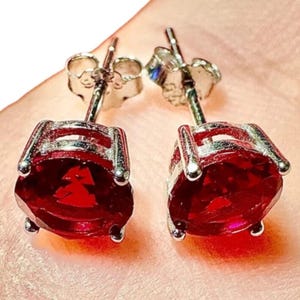 May include: A pair of stud earrings featuring vibrant, round, deep red gemstones set in a silver-colored metal. The earrings have a classic design with a post and butterfly backing, perfect for everyday wear.