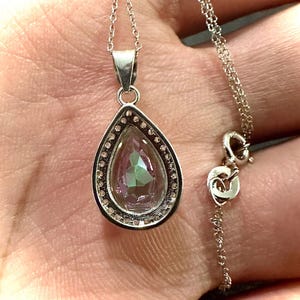 Pu&ograve; includere: Collana d'argento con un ciondolo a forma di goccia. Il ciondolo ha una gemma iridescente, circondata da piccole pietre trasparenti. La delicata catena d'argento &egrave; visibile. Il ciondolo misura circa 2 cm.