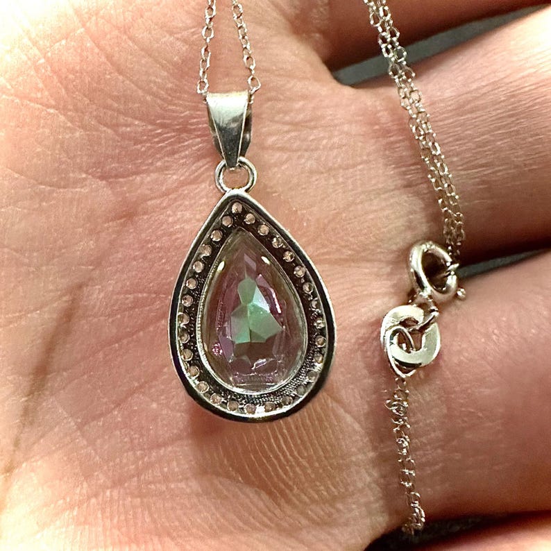 Pu&ograve; includere: Collana d'argento con un ciondolo a forma di goccia. Il ciondolo ha una gemma iridescente, circondata da piccole pietre trasparenti. La delicata catena d'argento &egrave; visibile. Il ciondolo misura circa 2 cm.