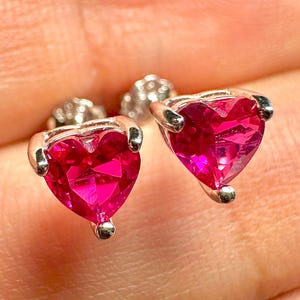 May include: A pair of heart-shaped stud earrings. Each earring features a vibrant, faceted, hot pink gemstone set in a silver-colored metal. The heart-shaped stones are the focal point, catching the light and creating a sparkling effect.
