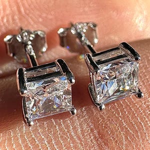 May include: A pair of square-cut diamond stud earrings. The earrings are set in a silver metal with a post and butterfly backing. The diamonds are clear and sparkling, reflecting light.