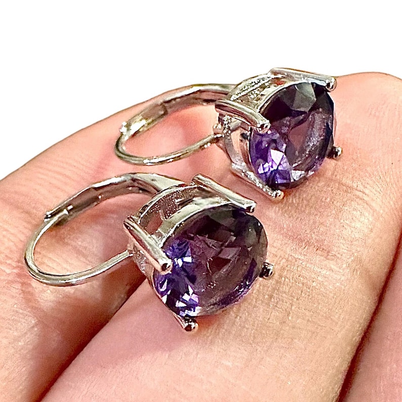 K&ouml;nnte beinhalten: Ein Paar silberfarbene Klappcreolen, jeweils mit einem gro&szlig;en, oval geschliffenen, tiefvioletten Edelstein. Die Edelsteine sind in einer Vier-Zacken-Fassung gefasst, die ihre Farbe und Brillanz hervorhebt.