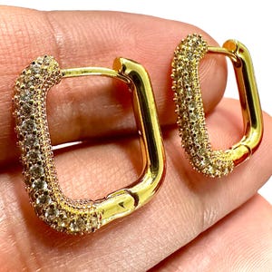 May include: A pair of gold-colored, square hoop earrings. One side of each earring is encrusted with small, clear stones. The earrings are held against a light-colored background.