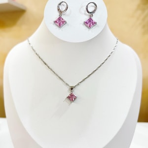 Könnte beinhalten: Ein silberfarbenes Schmuckset mit Halskette und passenden Ohrringen. Die Halskette hat eine zarte Kette mit einem quadratischen rosa Edelsteinanhänger. Die Ohrringe sind im Creolen-Stil mit quadratischen rosa Edelsteinen. Das Set wird auf einer weißen Schaufensterpuppe präsentiert.