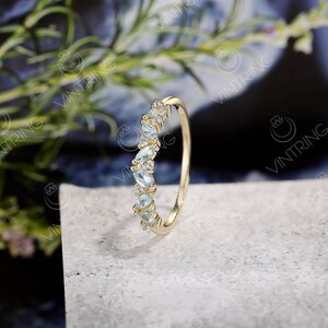 May include: A delicate gold band ring with a curved row of light blue marquise-shaped gemstones. The ring is set on a white textured surface.