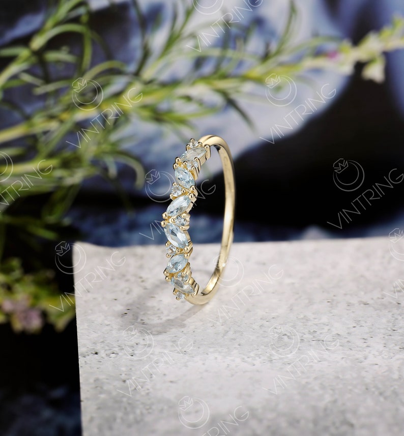 May include: A delicate gold band ring with a curved row of light blue marquise-shaped gemstones. The ring is set on a white textured surface.