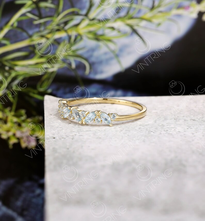 May include: A delicate gold band ring with a row of small, light blue gemstones. The ring is set on a white surface.