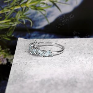 May include: A delicate silver ring with a curved row of light blue gemstones. The ring is set on a gray textured surface.