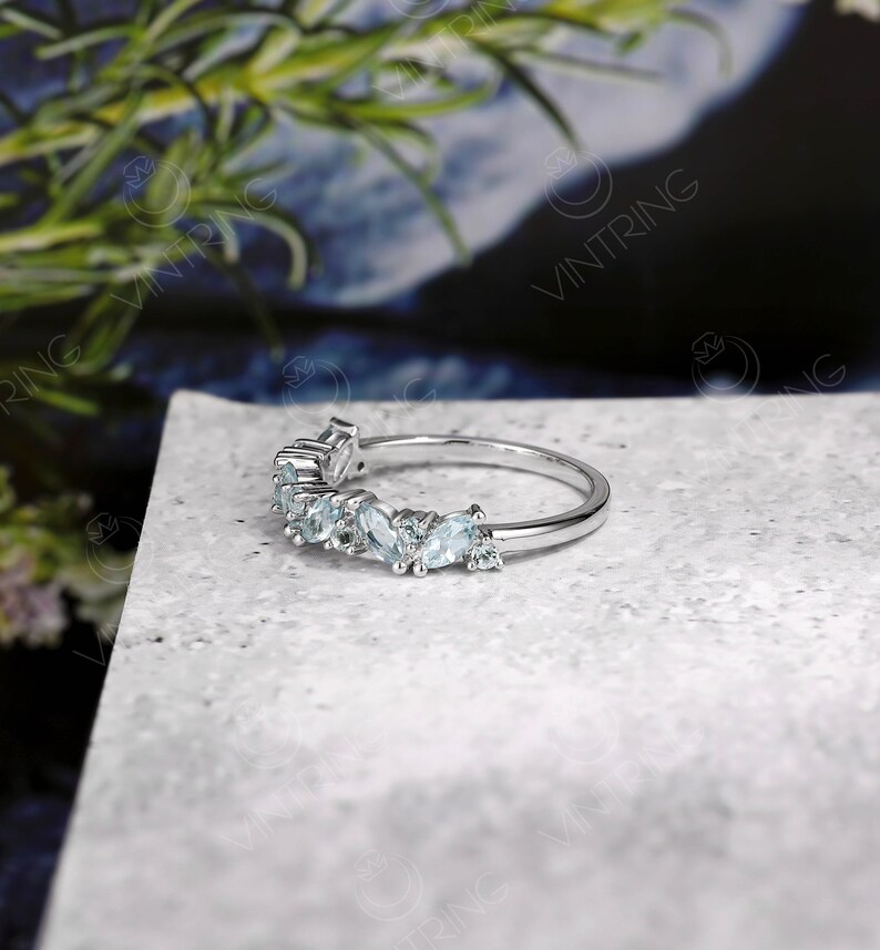 May include: A delicate silver ring with a curved row of light blue gemstones. The ring is set on a gray textured surface.