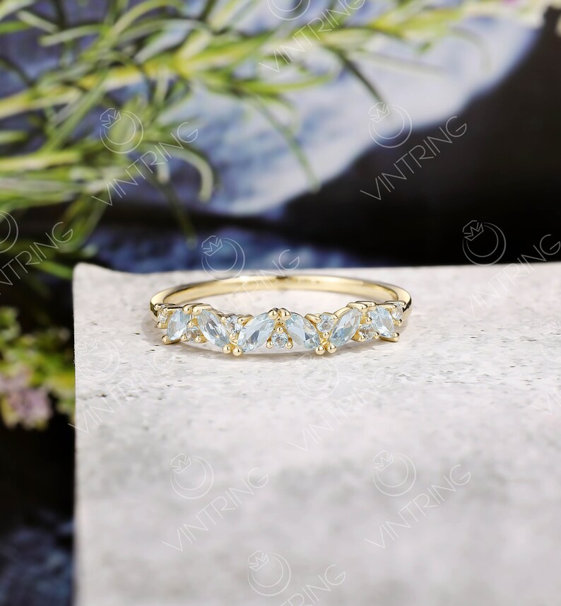 May include: A delicate gold band ring with a row of marquise-shaped aquamarine gemstones. The ring is set on a white background.