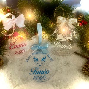 Pode incluir: Três enfeites de Natal circulares em acrílico transparente com fitas. Um tem um laço rosa e o nome Emma, um tem um laço azul claro e o nome Timéo com um desenho de rena, e um tem um laço dourado com o nome Léandro e um trenó de Papai Noel.