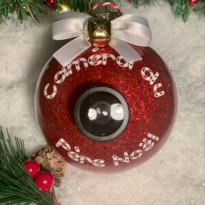 Peut inclure: Boule de Noël rouge pailletée avec un objectif d'appareil photo et les mots "Camera du Pere Noel" en blanc. L'ornement a un ruban blanc et des accents dorés. Décoration de vacances festive.