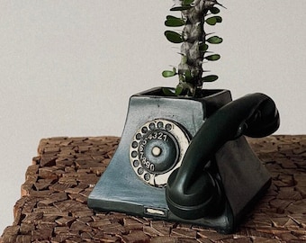 May include: A vintage-style rotary phone planter with a green finish and a black rotary dial. A small cactus plant is growing in the planter. The planter is sitting on a wooden surface.