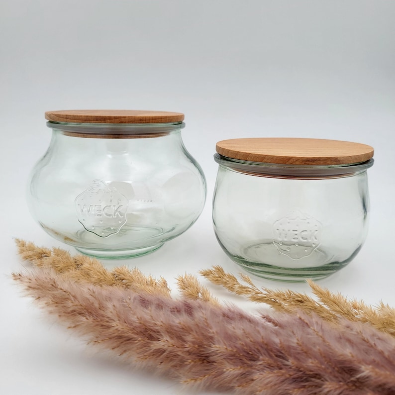 May include: Two clear glass jars with wooden lids. The jars have a rounded shape and the word "WECK" is embossed on the front. The lids are made of light-colored wood. Dried ornamental grass is in the foreground.