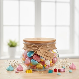 May include: A clear glass jar filled with colorful candies, including pink, blue, and red sweets. The jar has a wooden lid and a burlap trim. A wooden tag with the words "S&uuml;&szlig;e Momente" hangs from the jar.