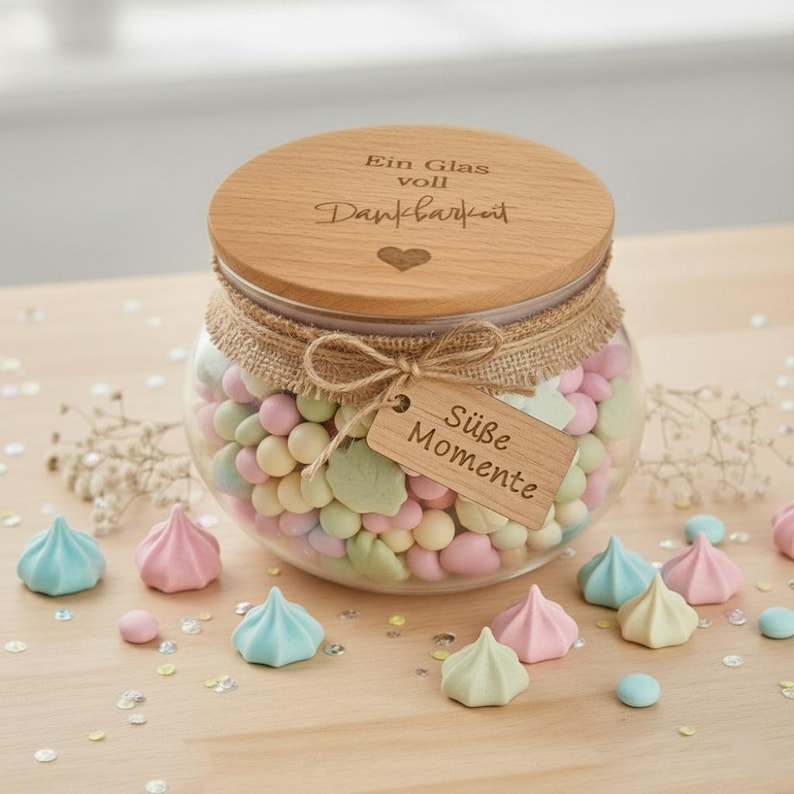May include: A glass jar filled with pastel-colored candies and a wooden lid. The lid is engraved with German text. A burlap ribbon and a wooden tag with the words "S&uuml;&szlig;e Momente" are tied around the jar. Several meringue cookies are scattered around the jar.