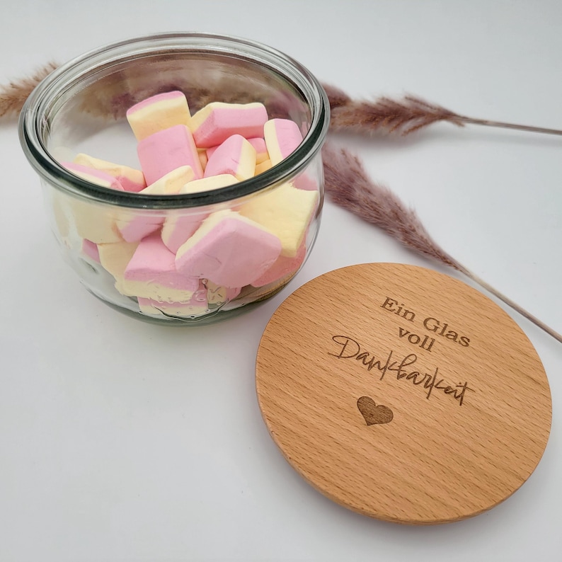 May include: A clear glass jar filled with pink and yellow marshmallows. A wooden lid with the text "Ein Glas voll Dankbarkeit" and a heart is next to the jar. The jar is on a white surface.