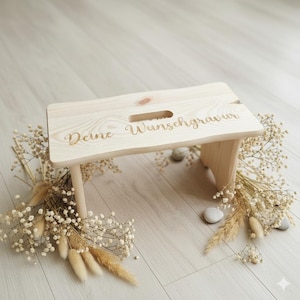 Könnte beinhalten: Ein helles Holztrittschemel mit dem eingravierten Text "Deine Wunschgravur". Der Hocker hat eine rechteckige Oberseite mit einem Griffausschnitt und vier Beinen. Getrocknete Blumen und kleine Steine umgeben den Hocker.