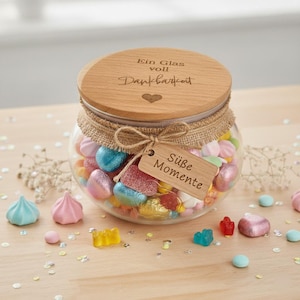 Ein Glas mit bunten Süßigkeiten, darunter blaue, rosa und gelbe Gummibärchen und herzförmige Süßigkeiten. Der Holzdeckel ist mit deutschem Text graviert. Ein Anhänger trägt die Aufschrift "Süße Momente".