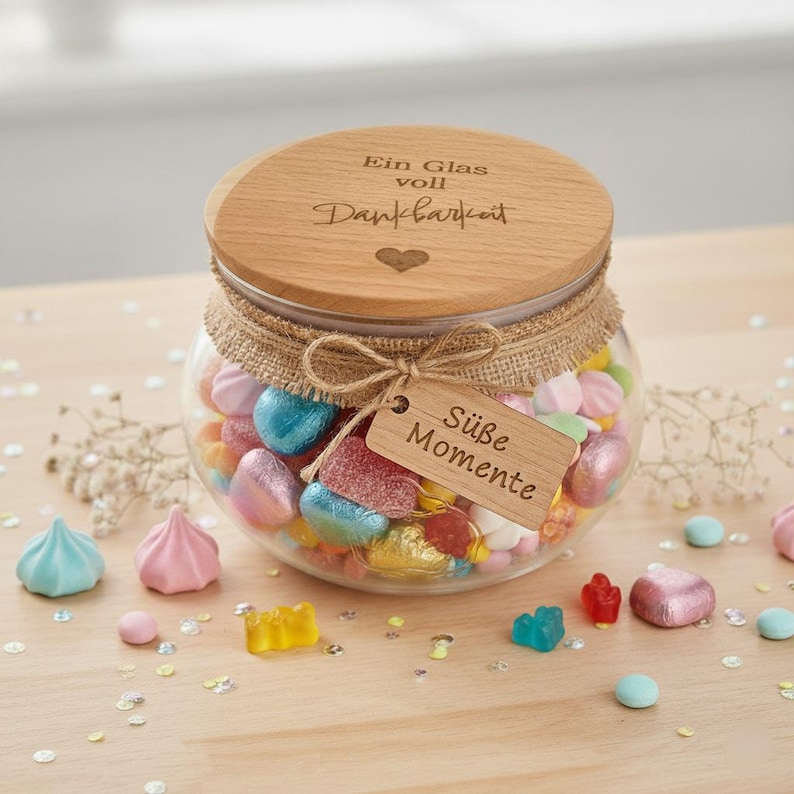 Ein Glas mit bunten Süßigkeiten, darunter blaue, rosa und gelbe Gummibärchen und herzförmige Süßigkeiten. Der Holzdeckel ist mit deutschem Text graviert. Ein Anhänger trägt die Aufschrift "Süße Momente".