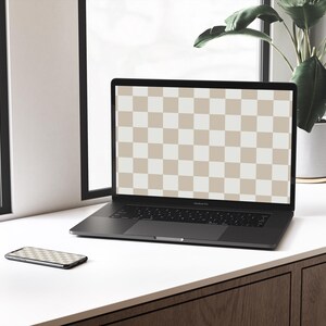 May include: A black laptop computer with a checkered screen is open on a white desk. A black smartphone is lying on the desk next to the laptop. A potted plant is visible in the background.