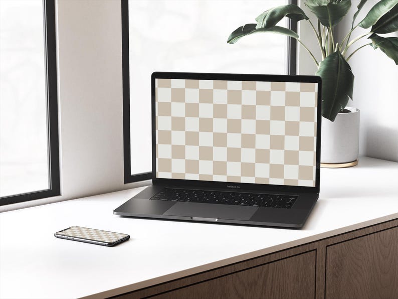 May include: A black laptop computer with a checkered screen is open on a white desk. A black smartphone is lying on the desk next to the laptop. A potted plant is visible in the background.