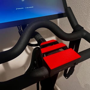 May include: Indoor exercise bike with a black frame and handlebars. The bike features a red foot platform and a screen displaying a user interface. The handlebars are curved and padded for comfort.