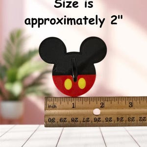 May include: A black, red, and yellow Mickey Mouse-shaped item. The item is approximately 2 inches in size. The text "Size is approximately 2" is above the item. A wooden ruler is below the item.