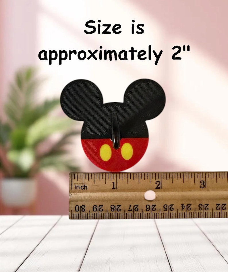 May include: A black, red, and yellow Mickey Mouse-shaped item. The item is approximately 2 inches in size. The text "Size is approximately 2" is above the item. A wooden ruler is below the item.