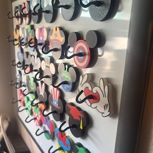May include: A collection of decorative wooden hooks in the shape of Mickey Mouse ears, each with a unique design. The hooks are mounted on a white board, with various colors and patterns, including polka dots, stripes, and character-themed designs.