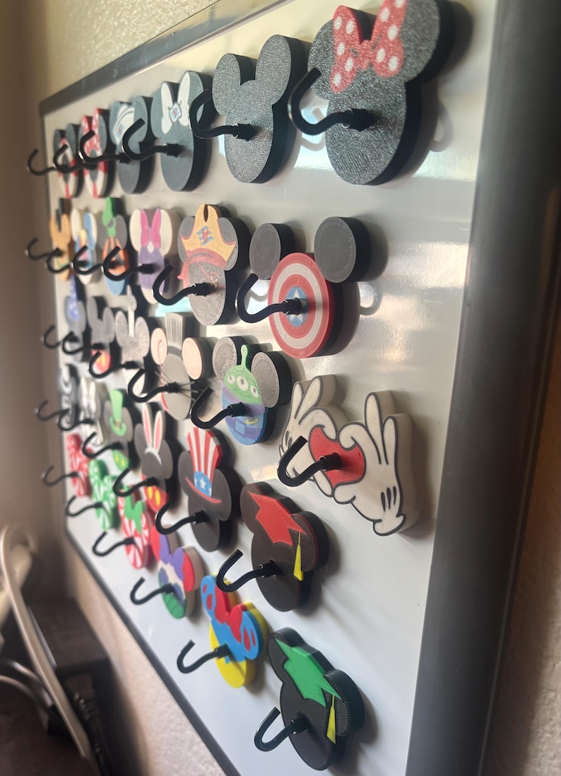 May include: A collection of decorative wooden hooks in the shape of Mickey Mouse ears, each with a unique design. The hooks are mounted on a white board, with various colors and patterns, including polka dots, stripes, and character-themed designs.