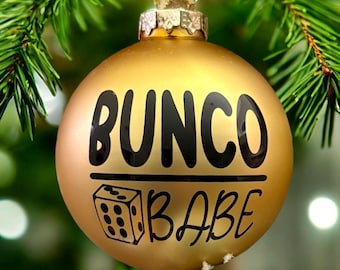 May include: A matte gold spherical Christmas ornament featuring the black text BUNCO above a horizontal line, with a graphic of a six-sided die and the black text BABE below the line. The ornament has a metallic silver cap at the top.