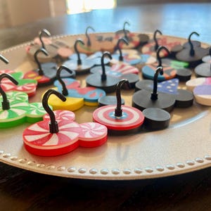 May include: A collection of decorative magnets in various designs, including Mickey Mouse shapes, candy cane patterns, and patriotic themes. Each magnet has a black hook attached, suitable for hanging small items. The magnets are arranged on a silver plate.