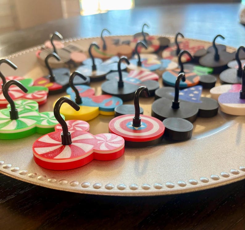 May include: A collection of decorative magnets in various designs, including Mickey Mouse shapes, candy cane patterns, and patriotic themes. Each magnet has a black hook attached, suitable for hanging small items. The magnets are arranged on a silver plate.
