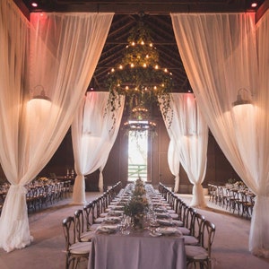 May include: A long, white-draped table with a grey tablecloth is set for a wedding reception in a rustic barn. The table is decorated with greenery and flowers. A large chandelier hangs above the table, and the barn is lit with string lights.