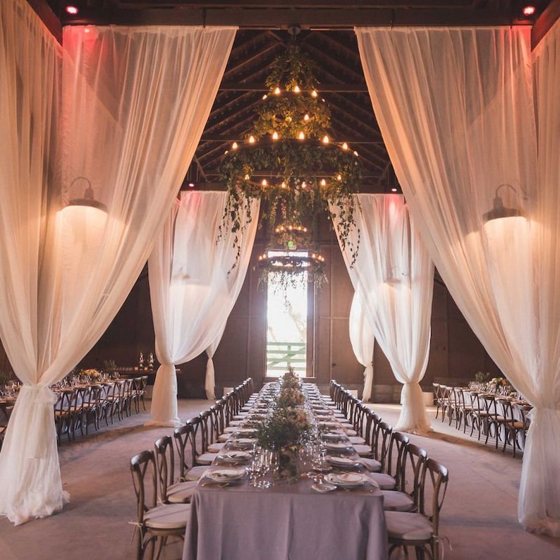 May include: A long, white-draped table with a grey tablecloth is set for a wedding reception in a rustic barn. The table is decorated with greenery and flowers. A large chandelier hangs above the table, and the barn is lit with string lights.