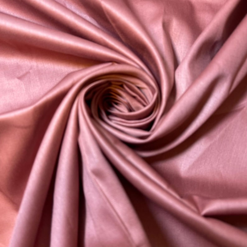 May include: Close-up of a soft, rose-colored fabric draped in a swirling pattern. The fabric has a subtle sheen and a smooth texture.