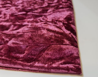 May include: Close-up of a folded piece of burgundy velvet fabric. The fabric has a crushed texture, creating highlights and shadows across its surface. The color is a deep, rich shade of red with a hint of purple.