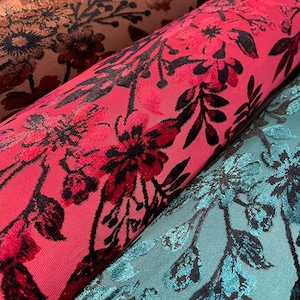 May include: Close-up of three rolls of floral patterned fabric. The first roll is a vibrant pink with dark red and black floral designs. The second roll is a teal colour with black floral designs. The third roll is a brown colour with black floral designs.