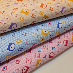 Può includere: Primo piano di tre rotoli di tessuto con un motivo ripetuto di orsetti dei cartoni animati, fiori e la scritta "I LOVE BEAR". Il tessuto è nei toni del giallo, blu e rosa, con uno sfondo a griglia.