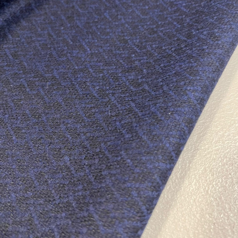 K&ouml;nnte beinhalten: Nahaufnahme eines gefalteten, marineblauen Stoffes mit einem dezenten, strukturierten Muster. Der Stoff hat eine dunkelblaue Basis mit einem helleren, eingewebten blauen Design. Der Rand des Stoffes liegt neben einer wei&szlig;en Oberfl&auml;che.