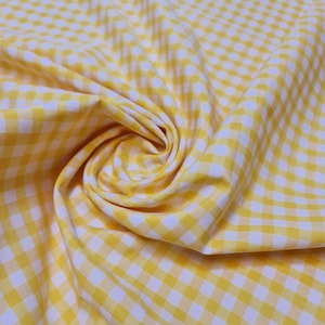 May include: Yellow and white gingham fabric with a checkered pattern. The fabric is folded and draped, showing the texture and weave.