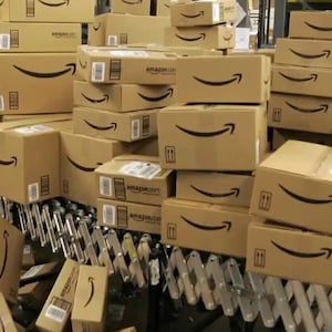 Może przedstawiać: Duży magazyn z wieloma brązowymi kartonami ułożonymi jeden na drugim. Pudełka są oznaczone logo Amazon i mają biały uśmiech. Pudełka są transportowane na taśmie przenośnikowej.