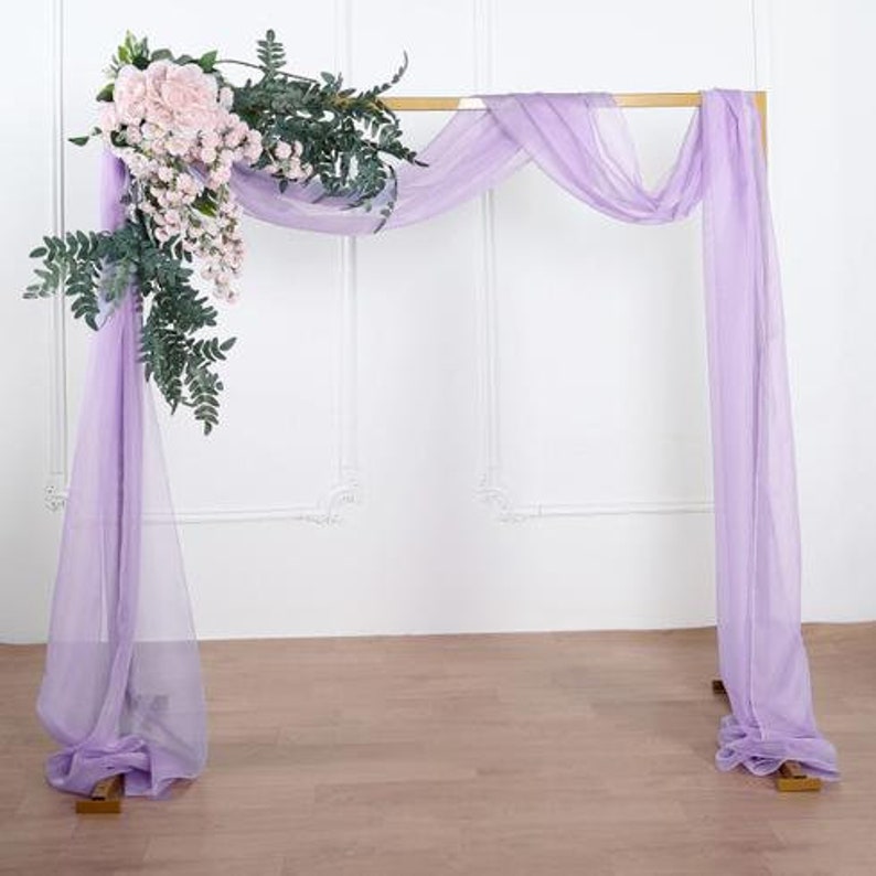 May include: A purple sheer fabric backdrop for a wedding ceremony or event. The fabric is draped over a gold metal archway and decorated with a bouquet of pink flowers.