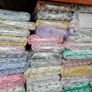 May include: A stack of fabric rolls wrapped in clear plastic. The fabric features various floral patterns in colors like blue, pink, yellow, and white. The rolls are neatly arranged on shelves, showcasing a variety of textile designs.