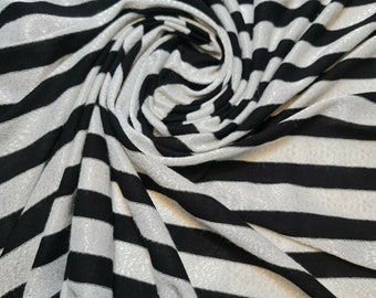 May include: Close-up of a fabric with a bold black and white striped pattern. The fabric is draped and swirled, creating a dynamic visual texture. The stripes are evenly spaced and run horizontally across the material.