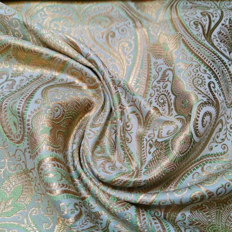 Op de afbeelding: Een close-up van een lichtblauwe stof met een paisley-patroon in goud en groen. De stof is zacht en luxueus, en het patroon is ingewikkeld en gedetailleerd.