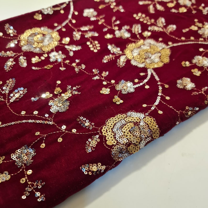 K&ouml;nnte beinhalten: Burgunderfarbener Samtstoff mit goldenen und silbernen Pailletten, die in einem Blumenmuster gestickt sind.
