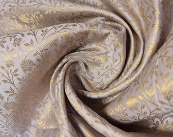 Op de afbeelding: Een close-up van een zachte, lichtbeige stof met een subtiel, herhalend patroon van gouden bloemmotieven. De stof is gedrapeerd en gevouwen, waardoor een textuur en een luxe uitstraling ontstaat.